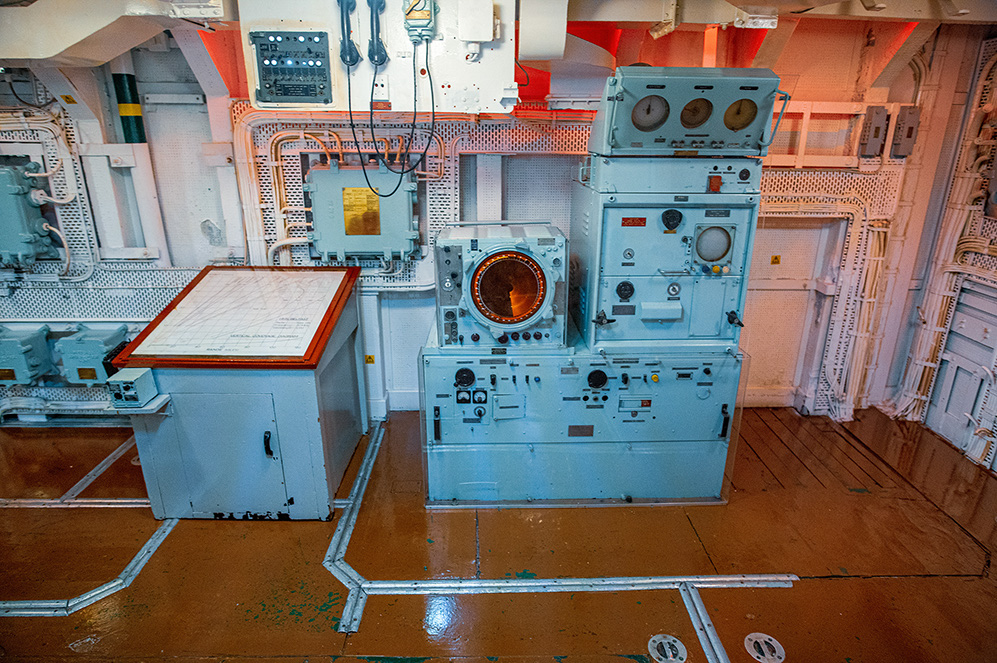 The image depicts an old, possibly vintage, control room or equipment room, likely from a ship or industrial setting. The room is filled with various pieces of equipment and control panels, all painted in a light blue color. The equipment includes dials, gauges, switches, and possibly some form of communication or radar equipment. The room has a somewhat worn and aged appearance, with visible signs of use and some rust. The floor is made of wood, and there are metal conduits running along it, connecting different pieces of equipment.