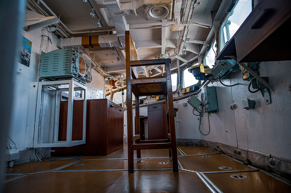 The image depicts the interior of a room, likely on a ship or a vessel. The room contains various equipment and machinery, with visible wiring and pipes along the walls and ceiling. There is a wooden chair in the center of the room, and the overall environment appears to be utilitarian and functional, designed for operational purposes.