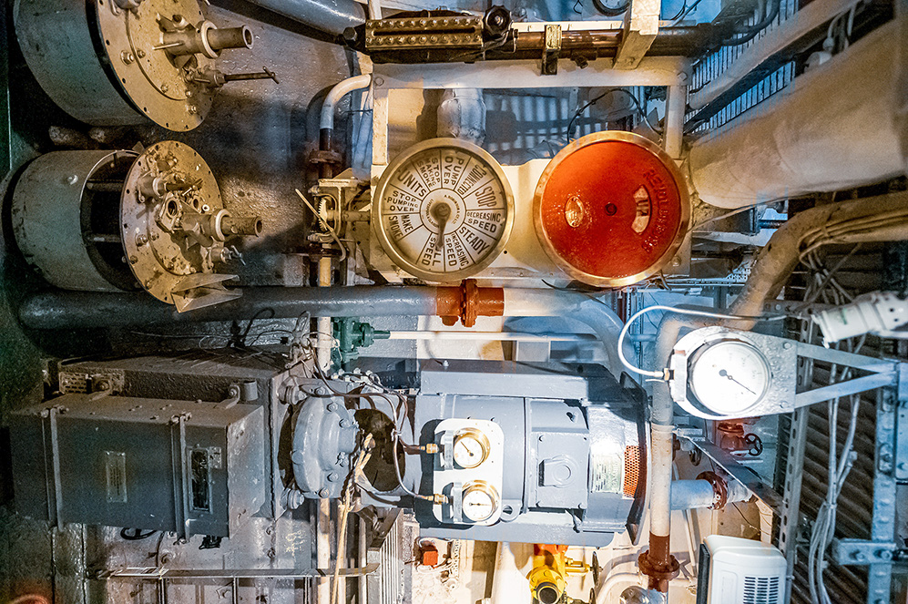The image depicts a complex and densely packed mechanical system, likely part of an industrial or maritime engine room. It features various gauges, valves, pipes, and machinery components, including a prominent red gauge and a large cylindrical object. The setup appears to be well-maintained, with visible labels and markings on the equipment, indicating a structured and organized environment. The overall scene suggests a highly technical and specialized area designed for monitoring and controlling mechanical operations.