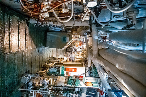 The image depicts the interior of the engine room, showcasing its complex and compact machinery and equipment. The view includes various pipes, wires, and mechanical components, highlighting the intricate and tightly packed nature of the vessel's systems. The lighting illuminates the narrow passage, revealing the detailed and industrial environment within the vessel.