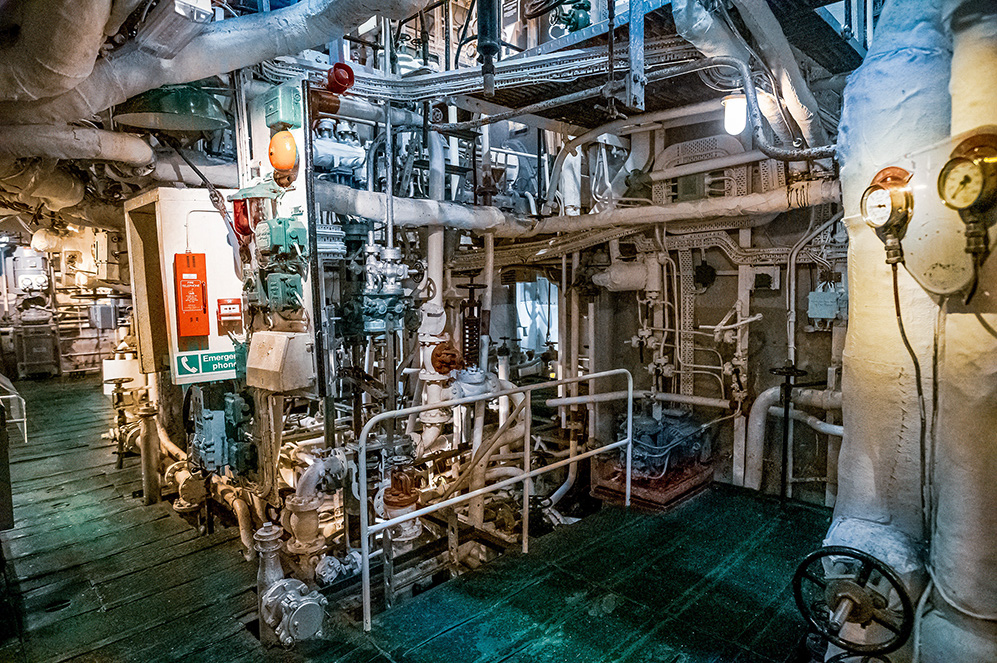 The image depicts an industrial setting, likely the engine room of a ship. It features a complex array of pipes, valves, gauges, and machinery. The environment appears to be well-lit with various lights and signs, including an emergency phone sign. The floor is covered with a greenish material, possibly to reduce slip hazards. The overall scene suggests a well-maintained and organized mechanical space.
