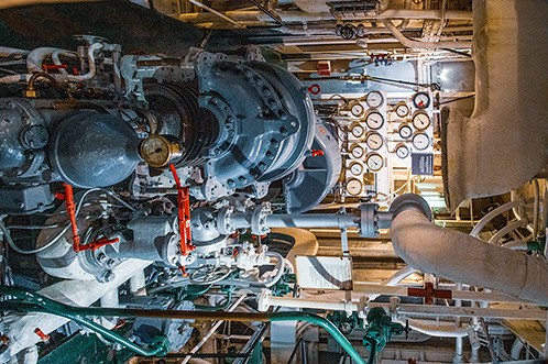 The image depicts an intricate and complex mechanical setup, likely an engine room or similar industrial environment. The machinery includes large, metallic components with various pipes, wires, and gauges. The area is well-lit, highlighting the detailed and meticulous arrangement of the equipment. The presence of a gloved hand suggests ongoing maintenance or inspection. Overall, the image conveys a sense of industrial precision and engineering complexity.
