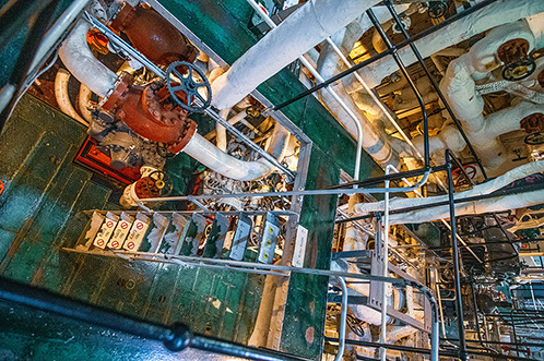 The image depicts an industrial or mechanical setting, likely the engine room of a ship or a similar vessel. It shows a complex array of machinery, pipes, and controls, with a prominent wheel and various gauges. The environment appears to be well-maintained and organized, with a focus on functionality and efficiency.