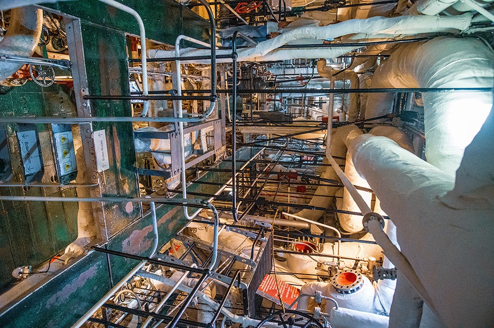 The image depicts the interior of an industrial facility, likely a power plant or a chemical processing plant. It shows a complex array of pipes, valves, and machinery, with some areas covered in protective insulation. The environment appears to be well-lit and organized, with safety railings and platforms for maintenance access. The overall scene suggests a highly technical and controlled industrial setting.