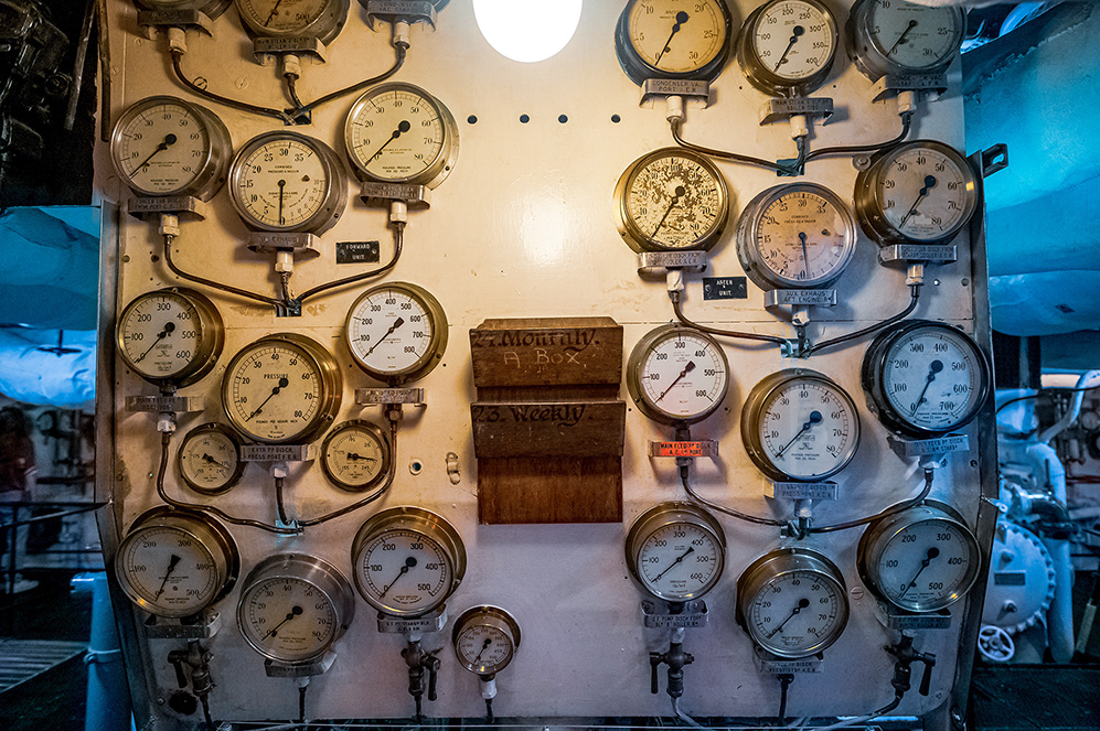 The image shows a control panel with numerous analog pressure gauges and valves, likely from an industrial or maritime setting. The gauges are arranged in a grid pattern and are connected by a network of pipes and tubes. There is a wooden box labeled 'Monthly' and 'Weekly' in the center of the panel. The overall appearance suggests an older, possibly vintage, mechanical system used for monitoring and controlling pressure in various systems.