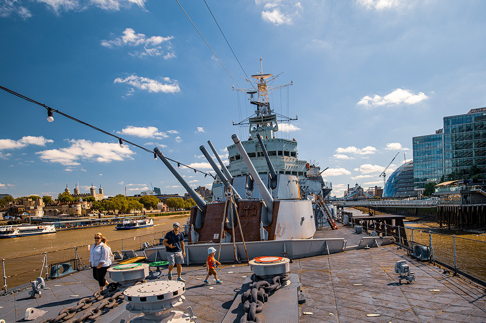 The image depicts a large naval ship docked at a harbor with several people walking on its deck. The ship is equipped with various antennas and equipment, and the background features a cityscape with modern buildings and historical structures, including a dome-shaped building and a bridge. The sky is clear with scattered clouds, and the overall scene suggests a sunny day at a busy waterfront.