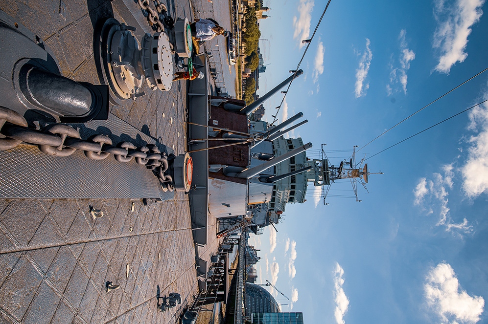 The image shows a large naval ship docked at a pier. The ship is equipped with various antennas and communication equipment, and its deck is visible with several features like lifebuoys and anchor chains. The sky is clear with some clouds, and the pier is made of stone with some people walking around. In the background, there are buildings and trees, indicating an urban setting near a body of water.