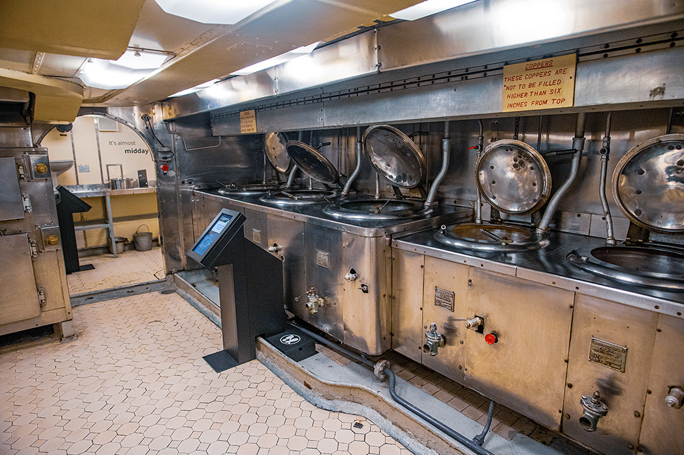 The image depicts a galley kitchen with large, industrial-sized cooking equipment, likely from a ship or large vessel. The equipment includes multiple copper pots with lids, and there is a sign indicating that the coppers should not be filled higher than six inches from the top. The floor is tiled, and there is a touchscreen display in the foreground, possibly for informational purposes. The overall setting appears to be well-maintained and organized.