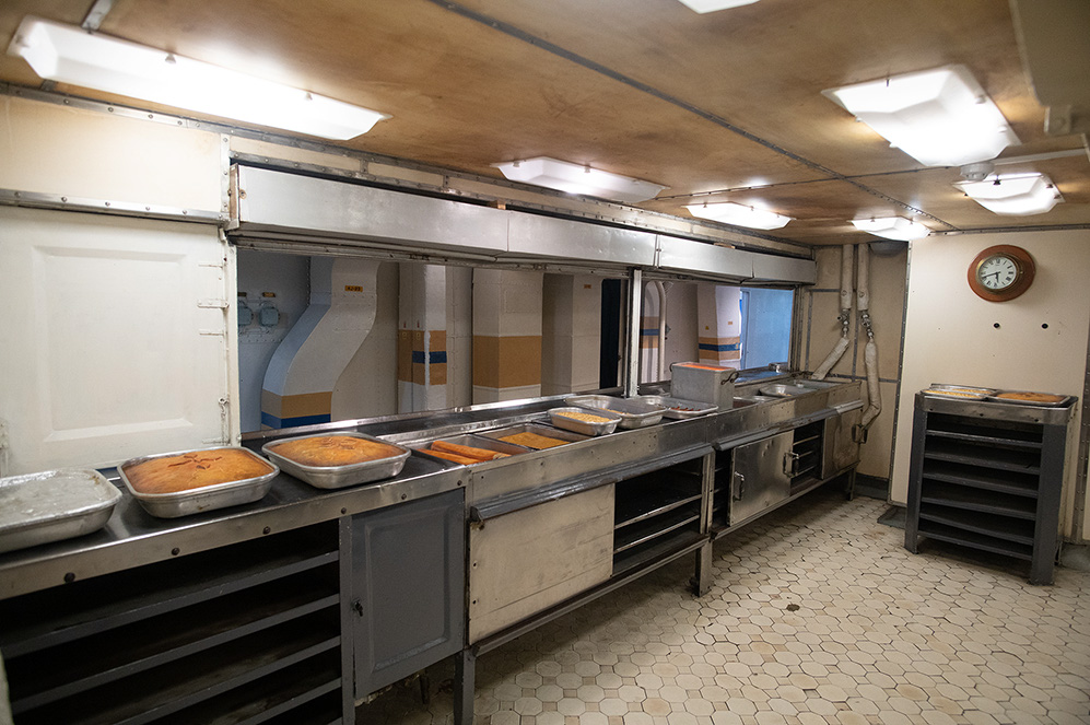 The image depicts a large industrial kitchen with stainless steel counters, trays of food, and various cooking equipment. The kitchen is well-lit with overhead lights and has a tiled floor. There is a clock on the wall, and the overall environment appears clean and organized.