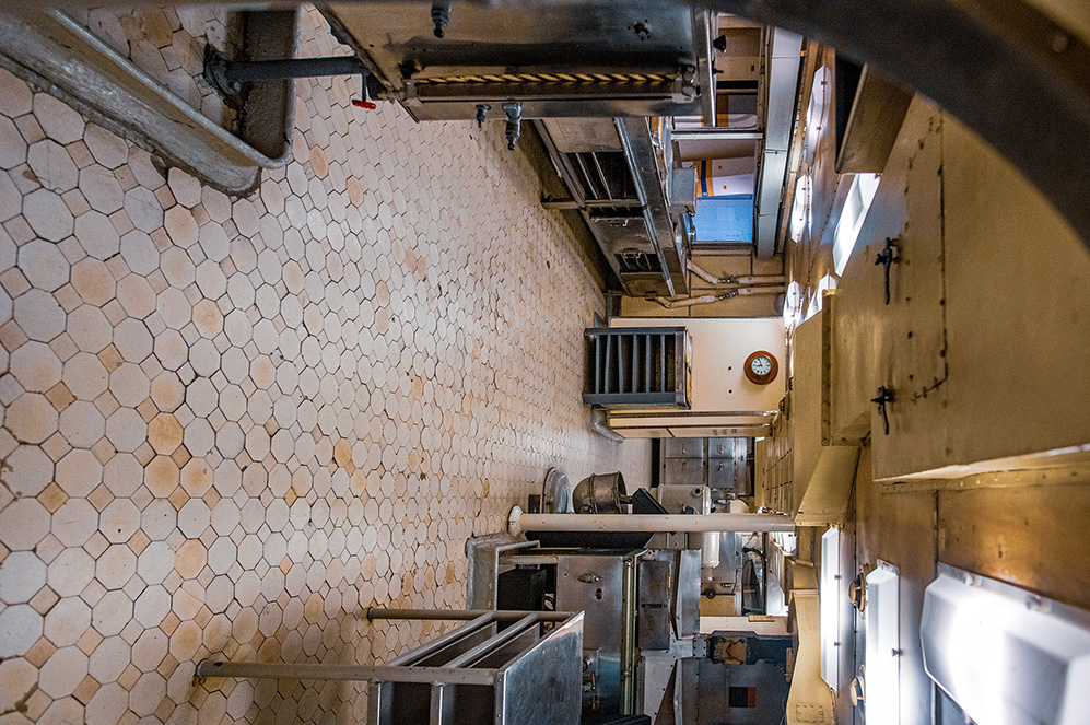 The image depicts an industrial or commercial kitchen with hexagonal tiled walls, stainless steel surfaces, and various kitchen equipment. The space appears to be well-lit and organized, with visible ventilation systems and a clock on the wall.