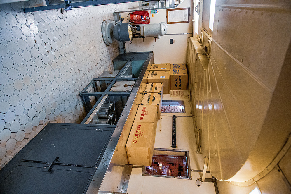 The image shows an interior space, likely a ship's galley or storage area. There are several cardboard boxes labeled with items such as 'Cup-a-Soup' and 'Instant Tea' stacked on a counter. The walls are covered with hexagonal white tiles, and there is a fire extinguisher mounted on the wall. Various pieces of equipment and storage compartments are visible, indicating a functional and utilitarian space.