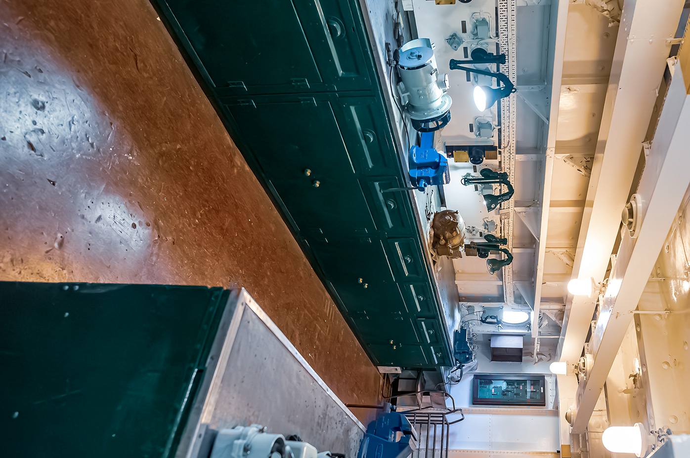 The image depicts a narrow corridor with a low ceiling, likely within an industrial or mechanical setting. The corridor is lined with green cabinets on one side and various mechanical components, pipes, and wires on the other. The ceiling has exposed beams and several lights, some of which are on. The floor appears to be metallic and slightly worn, with visible signs of use and some water or liquid spillage.