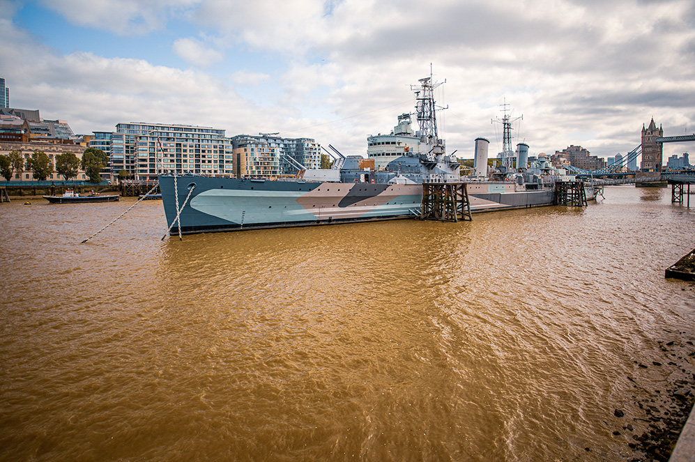 The image shows a large, camouflage-painted naval ship docked on the River Thames in London. The ship is moored near modern buildings and the iconic Tower Bridge, which is visible in the background. The sky is partly cloudy, and the water of the river appears brown and murky. The ship appears to be a museum or historical exhibit, as it is secured to the shore with wooden supports.