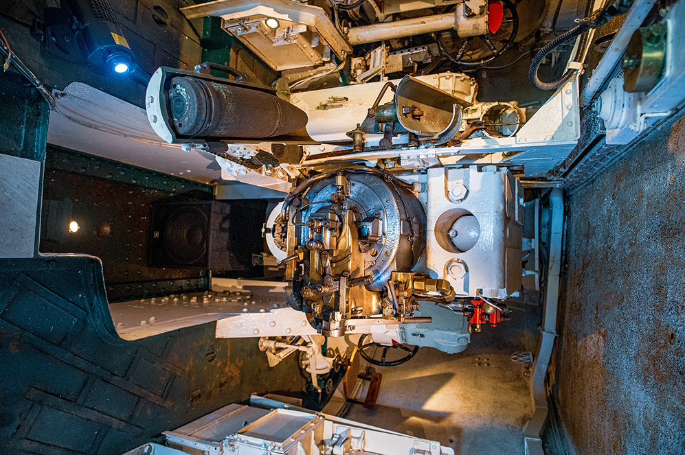 The image depicts the interior of a military tank, showing the turret mechanism and main gun. The view is from above, looking down into the turret, revealing various mechanical components and the complex structure of the tank's armament system.