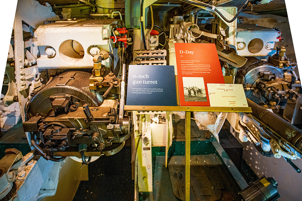 The image shows the interior of a military vessel, specifically focusing on a 6-inch gun turret. There are informational placards describing the turret and its historical context, including its use during D-Day. The turret is part of a museum exhibit, providing details about its operation and significance in naval history.