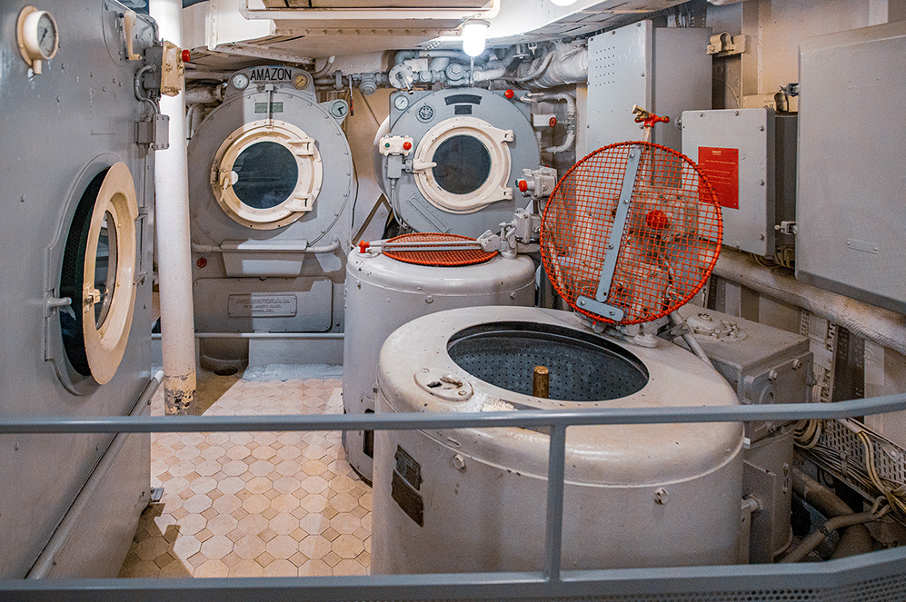 The image shows an industrial laundry room with large washing machines and dryers, likely on a ship or in a large facility. The equipment appears to be old and well-used, with a fan placed in front of one of the machines, possibly for drying clothes. The room is enclosed with a glass barrier, and the floor is covered with small, light-colored tiles.