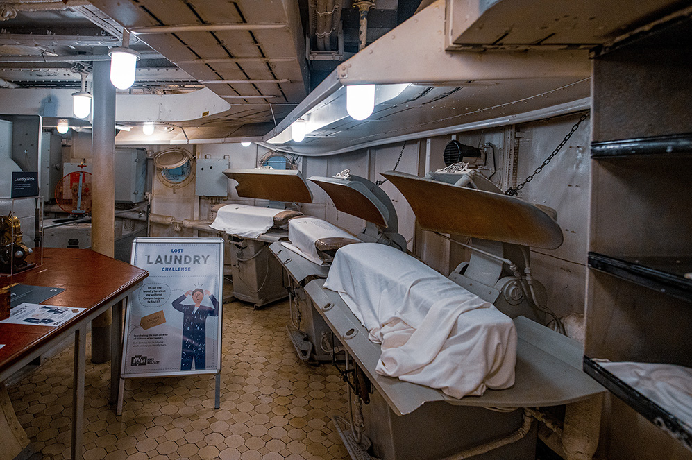 The image depicts an old-fashioned laundry room, likely on a ship, with multiple mangle irons and a sign promoting a 'Laundry Challenge'. The room is equipped with vintage machinery and has a rustic, utilitarian appearance. The sign suggests an interactive exhibit, inviting visitors to participate in a historical laundry task.