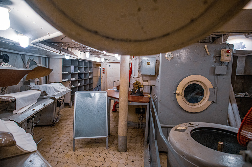 The image depicts an interior space, likely a medical or examination room on a ship or vessel. The room features several medical beds with white covers, shelves for storage, a metal cabinet, and a porthole door. The floor is tiled with small, hexagonal tiles, and the ceiling has a curved, metallic surface with lights.