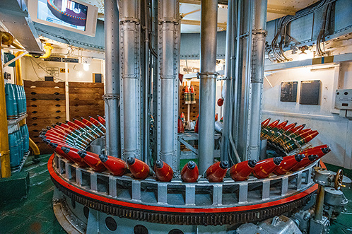 The image shows an industrial setting with a large, circular machine that has multiple vertical pipes and red-tipped components. The machine appears to be part of a manufacturing or processing facility, with various other equipment and structures visible in the background.