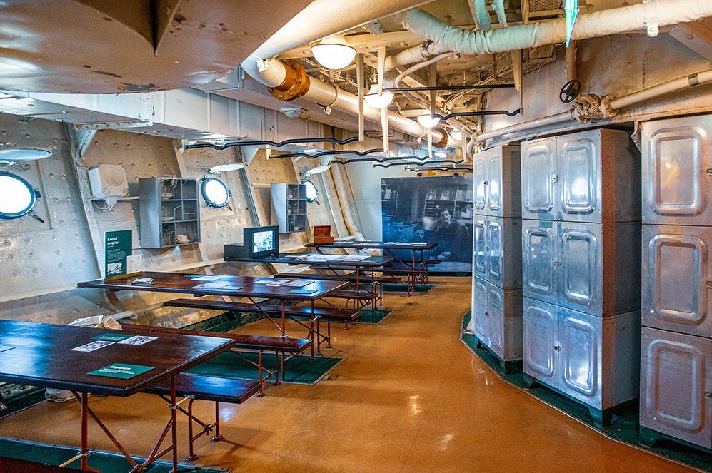 The image depicts the interior of a historical ship, likely a naval vessel, showcasing a mess hall or dining area. The room features several long wooden tables and benches, with round porthole windows along the wall. The walls and ceiling have exposed pipes and industrial elements, giving it a utilitarian and vintage appearance. There are also large metal cabinets and a television screen displaying information, suggesting the space is part of a museum or exhibit.