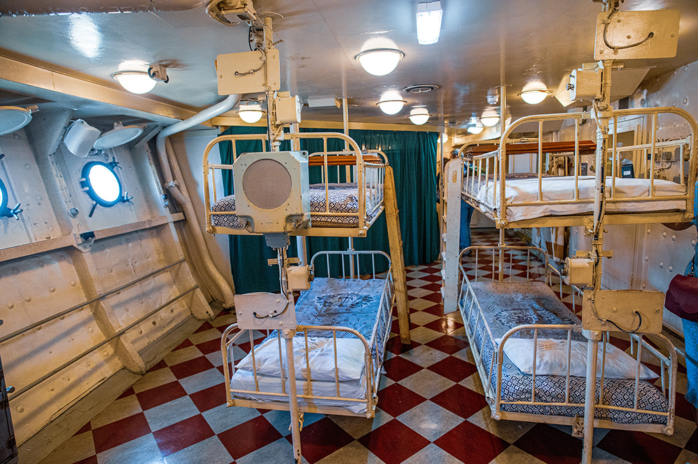 The image depicts a small, compact room with multiple bunk beds, likely a dormitory or shared sleeping quarters on a ship or vessel. The room features metal walls with porthole windows, a checkered floor, and a green curtain dividing the space. The bunk beds are arranged in a way to maximize the use of limited space, with some beds stacked three levels high. The overall design suggests a utilitarian and functional environment, prioritizing efficiency and practicality