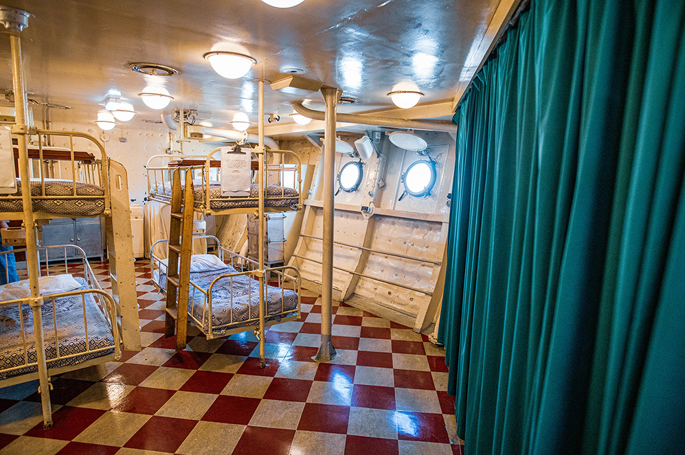 The image depicts the interior of a ship's cabin with multiple bunk beds arranged in a compact space. The cabin features a checkered floor, metal bunk beds with mattresses, and portholes for natural light. The ceiling is adorned with circular light fixtures, and a green curtain serves as a partition. The overall setting suggests a utilitarian and functional design typical of maritime accommodations.