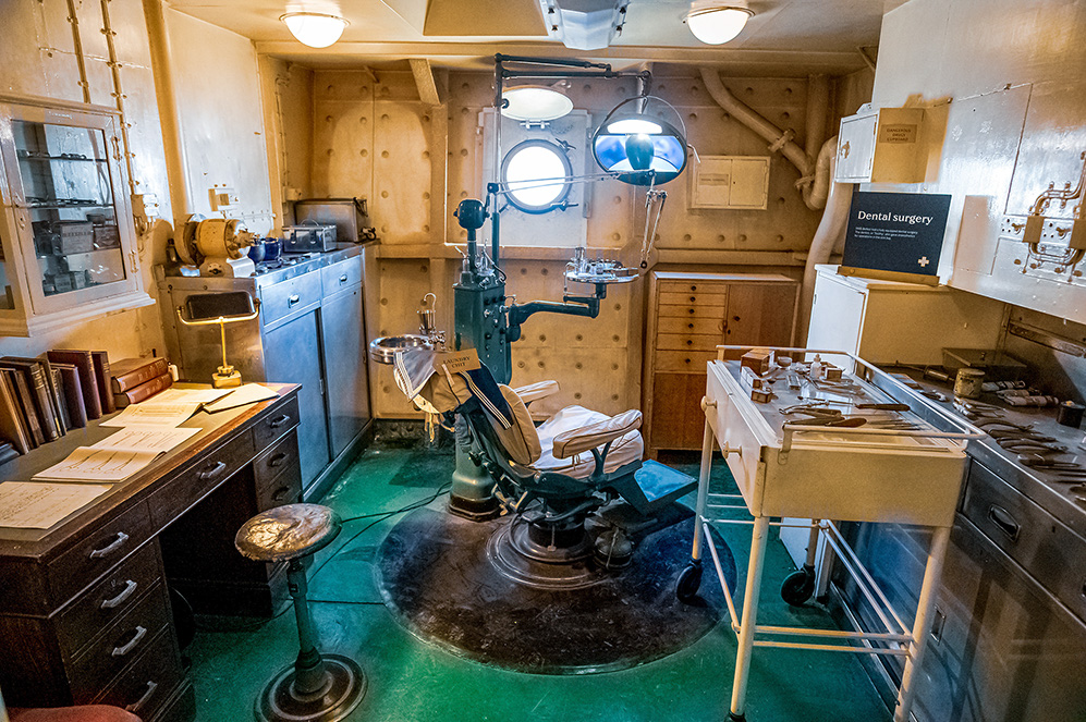 The image depicts an old-fashioned dental surgery room with vintage equipment, including a dental chair, various tools, and cabinets. The room has a historical feel, with a green-tinted floor and round porthole windows, suggesting it might be on a ship. There are books, instruments, and other medical supplies scattered around, giving it an authentic, well-used appearance. A sign on the wall reads 'Dental surgery,' confirming the room's purpose. The overall ambiance is one of historical preservation, likely part of a museum exhibit.