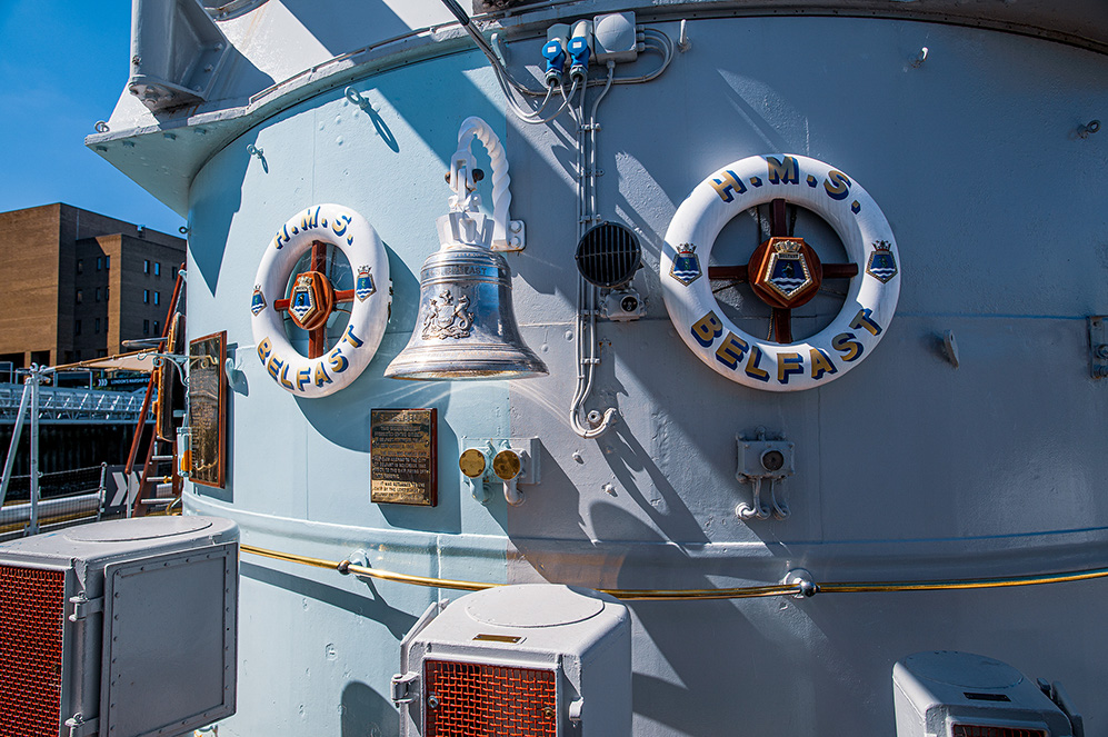 The image shows a close-up view of the side of a ship named H.M.S. Belfast. The ship's name is prominently displayed on two lifebuoys. There is a large, ornate bell mounted on the ship's side, and several plaques and insignias are visible, indicating the ship's identity and possibly its achievements or history. The ship appears to be docked, with some buildings visible in the background.