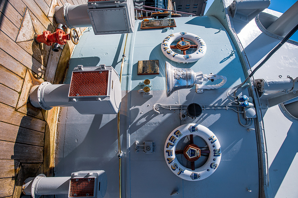 The image shows the deck of a ship, specifically the USS Iowa Museum, as indicated by the lifebuoys with 'USS Iowa' inscribed on them. The deck features various equipment, including large gray pipes, a red fire hydrant, and a white box with a grid pattern. There is also a plaque and a sculpture of an eagle, adding to the historical and maritime ambiance of the scene.