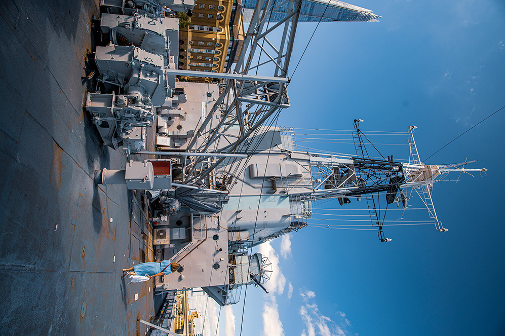 The image shows an aerial view of a naval ship docked at a port. The ship is equipped with various antennas and communication equipment, and a tall structure, possibly a mast or radar tower, rises from its deck. The surrounding area includes buildings and a clear blue sky with a few clouds. The ship appears to be in a well-maintained condition, indicating it is either in active service or undergoing maintenance