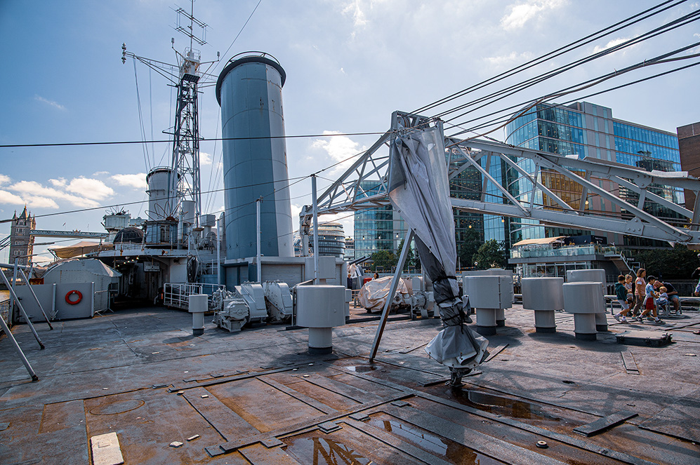 The image depicts the deck of a large ship, likely a museum ship given the modern buildings in the background and the presence of visitors. The deck features various maritime equipment, including a large crane covered with a tarp, several cylindrical structures, and a complex network of wires and rigging. The ship's deck is made of metal plates, some of which appear rusted. The background shows contemporary urban buildings, suggesting the ship is docked in a city setting.