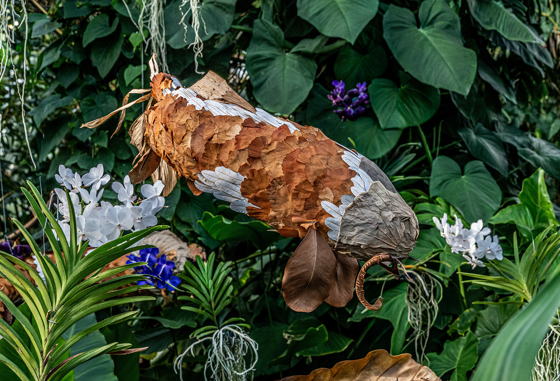 The image depicts a decorative sculpture of a fish crafted from natural materials such as leaves and branches. The fish is integrated into a lush, green environment with various plants, including white and purple flowers. The sculpture is detailed, with the bird appearing to have wings and feathers made from different types of leaves and bark. The overall scene is vibrant and natural, blending art with nature seamlessly.