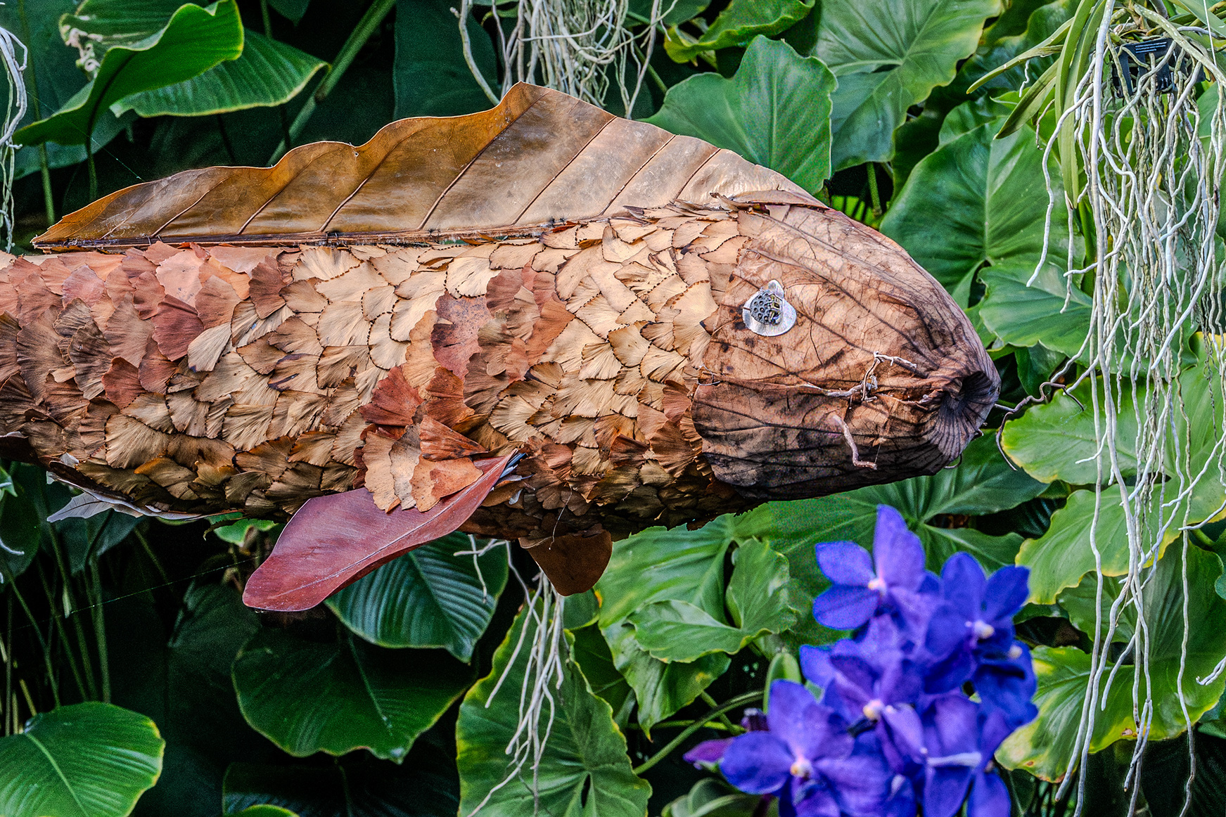 The image depicts a fish sculpture crafted from natural materials such as leaves and bark, placed among green foliage and purple flowers. The sculpture is detailed and realistic, blending seamlessly with its surroundings. The fish appears to be swimming among the plants, creating a harmonious and artistic scene.