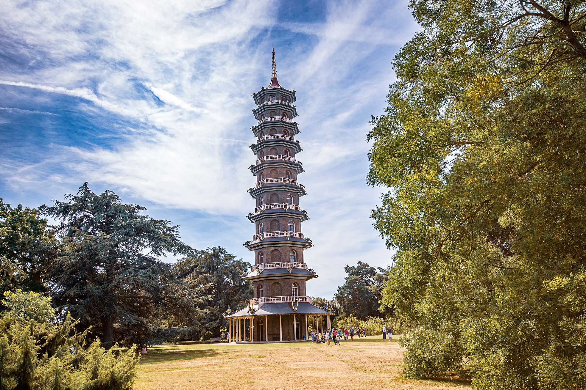The image depicts a tall, multi-tiered pagoda-style tower situated in a lush, green park. The tower has several levels with balconies and is topped with a spire. The surrounding area is filled with trees and people walking around, suggesting it is a popular spot for visitors.