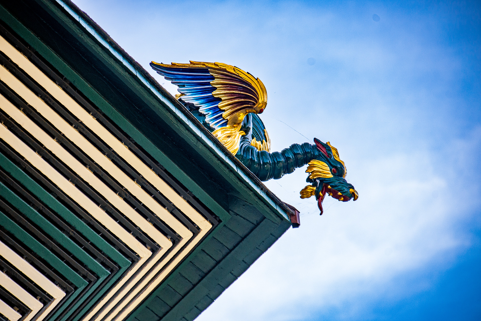 The image depicts a colorful, ornate sculpture of a winged dragon figure mounted on the corner of a building. The figure has a blue body with golden wings and is holding a bunch of grapes. The building has a green and white striped pattern.