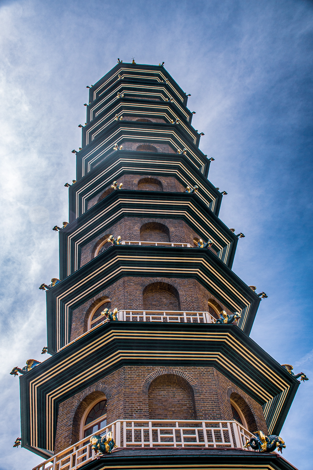 The image depicts a tall, multi-tiered pagoda-style tower with ornate decorations. The tower is constructed from brick and features multiple levels, each with arched openings and detailed cornices. The structure is adorned with golden ornamental elements, including statues of mythical creatures at each corner of the eaves. The sky in the background is clear with some clouds, providing a stark contrast to the intricate details of the tower.