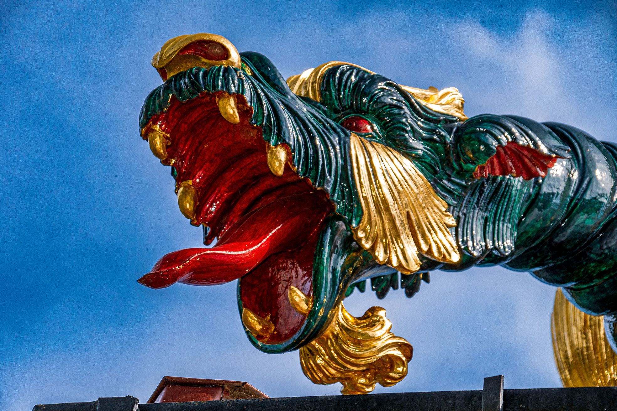 The image depicts a colorful and intricate sculpture of a dragon's head, featuring vibrant colors such as red, green, gold, and black. The dragon has detailed features including a red tongue, gold teeth, and a green body with gold accents. The sculpture is set against a clear blue sky, suggesting it is located outdoors, possibly on a building or temple.