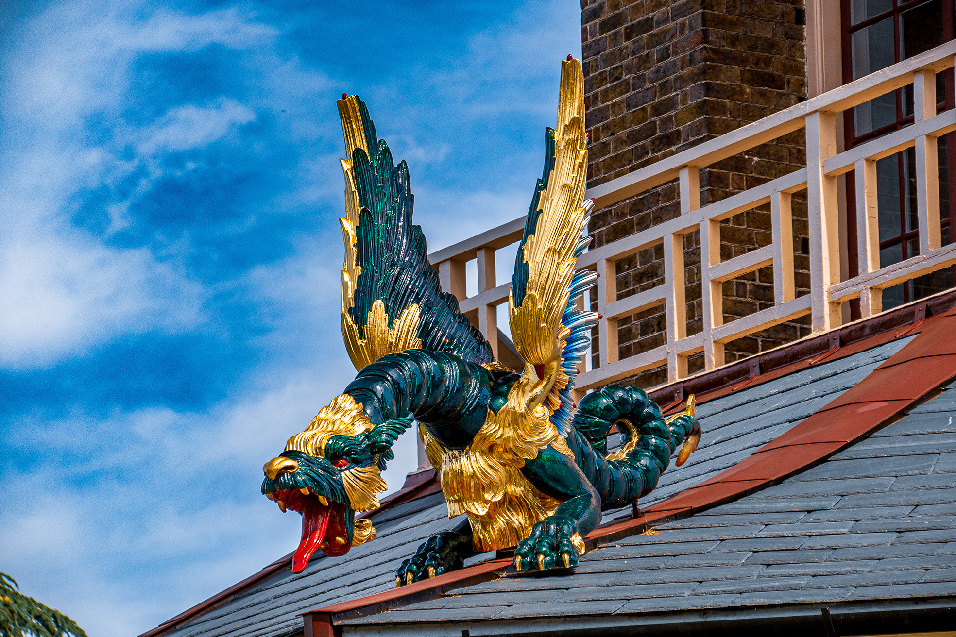 The image depicts a decorative dragon figure on the roof of a building. The dragon is intricately designed with vibrant colors, predominantly green and gold, and features detailed scales and an open mouth. The building has a traditional architectural style with a tiled roof and white railings. The sky in the background is clear with a few clouds.