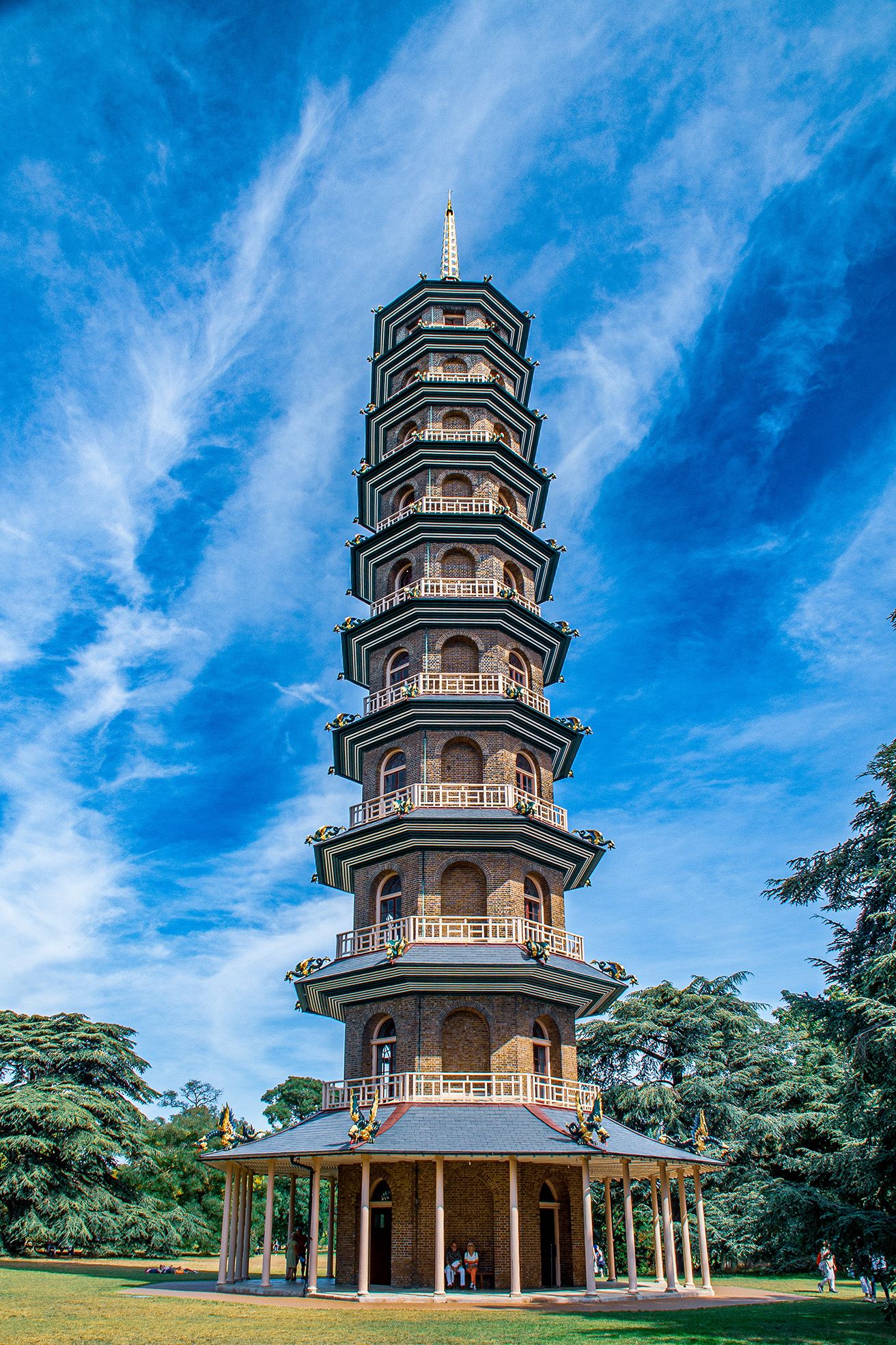 The image depicts a tall, multi-tiered pagoda-style tower situated in a lush, green park. The tower features several levels with arched windows and balconies, topped with a spire. The structure is surrounded by trees and a clear blue sky with some wispy clouds. There are a few people standing at the base of the tower, and the overall scene appears to be a peaceful, well-maintained park area.