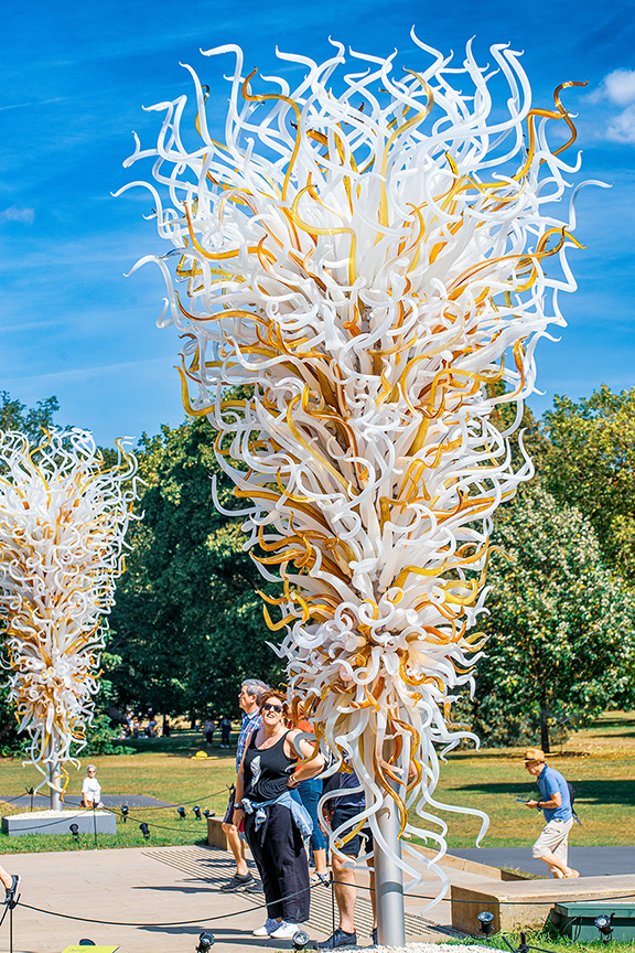 The image depicts a vibrant outdoor scene featuring large, intricate glass sculptures resembling trees with white and yellow tendrils. The sculptures are set in a park with people walking and enjoying the surroundings on a sunny day.