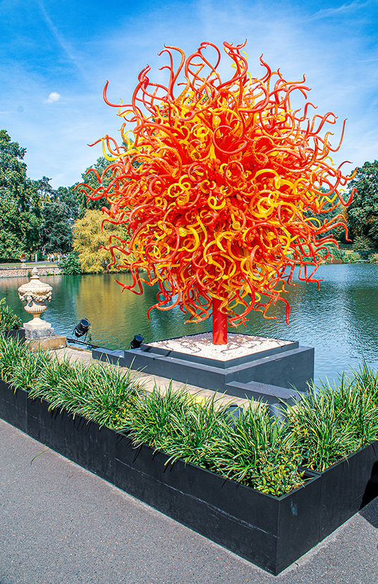 The image depicts a vibrant, colorful glass sculpture situated by a pond in a park. The sculpture, resembling a tree with red and yellow tendrils, stands on a square base. The surrounding area includes a neatly trimmed grass border and a pathway. The scene is set against a backdrop of trees and a clear blue sky.