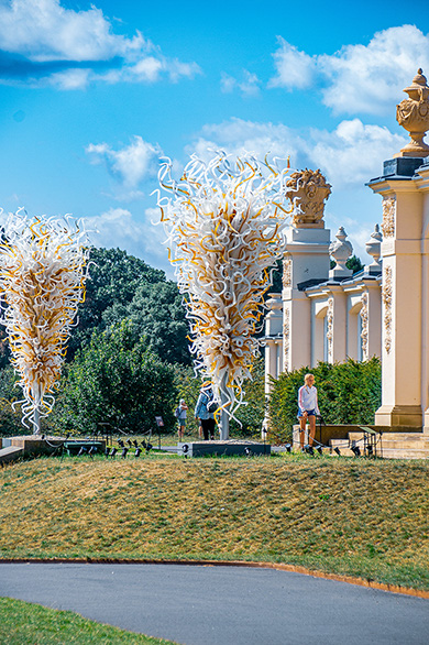 The image depicts an outdoor scene with large, abstract, golden sculptures resembling trees or bushes with intricate, swirling patterns. The sculptures are situated in a well-maintained park or garden area with a manicured lawn and a paved path. In the background, there is a classical-style building with columns and ornate decorations. A person is seen sitting on a bench, enjoying the view. The sky is clear with a few scattered clouds, indicating a bright and sunny day.