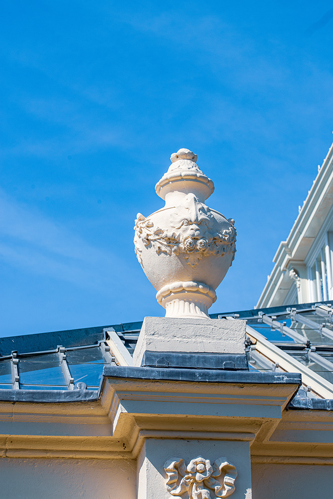 The image shows an ornate architectural detail on the top of a building, featuring a decorative urn or vase with intricate designs and a finial on top, set against a clear blue sky. The building has a light-colored facade with a balcony and decorative elements.