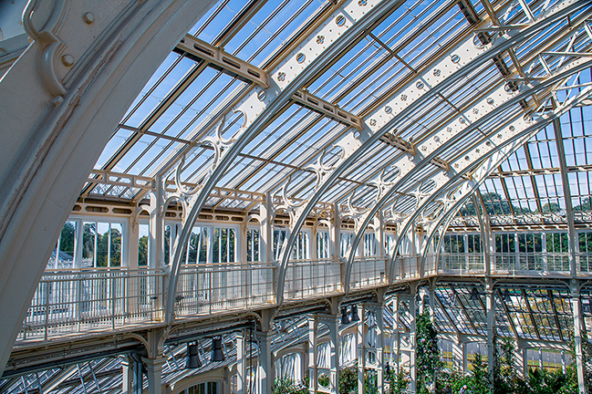 The image shows an ornate architectural detail on the top of a building, featuring a decorative urn or vase with intricate designs and a finial on top, set against a clear blue sky. The building has a light-colored facade with a balcony and decorative elements.