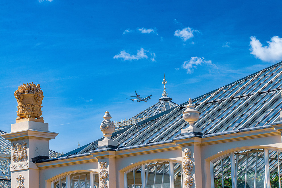The image depicts a clear blue sky with a few scattered clouds. In the foreground, there is a building with ornate architectural details, including decorative columns and a crest. The building has a glass roof, and in the background, an airplane is captured mid-flight, ascending into the sky.