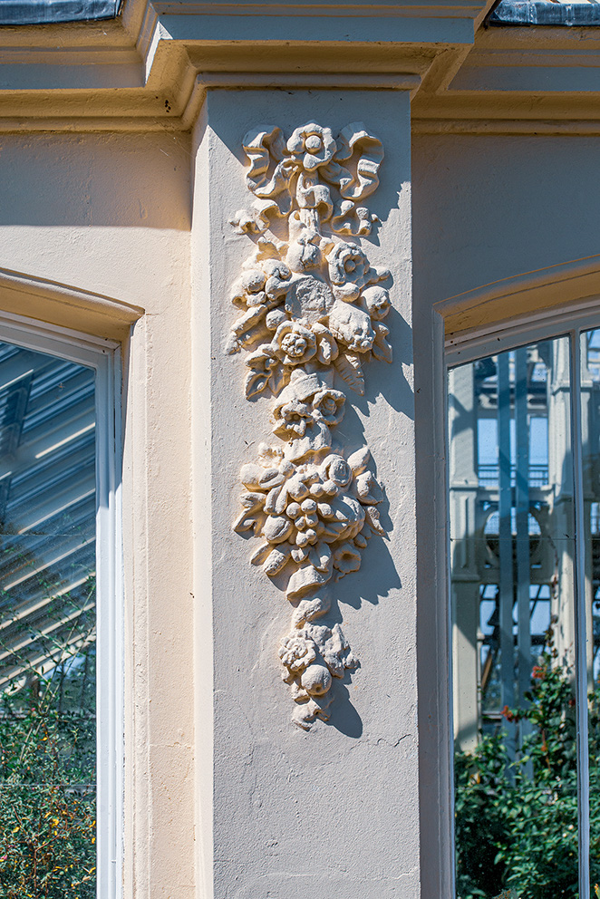 The image showcases an ornate architectural detail on a building. The detail features a vertical arrangement of sculpted floral and shell-like elements, set within a niche on a light-colored wall. The surrounding structure includes windows with white frames and a decorative balcony with a metal railing. The scene is bathed in natural light, highlighting the intricate design and the texture of the sculpted elements.