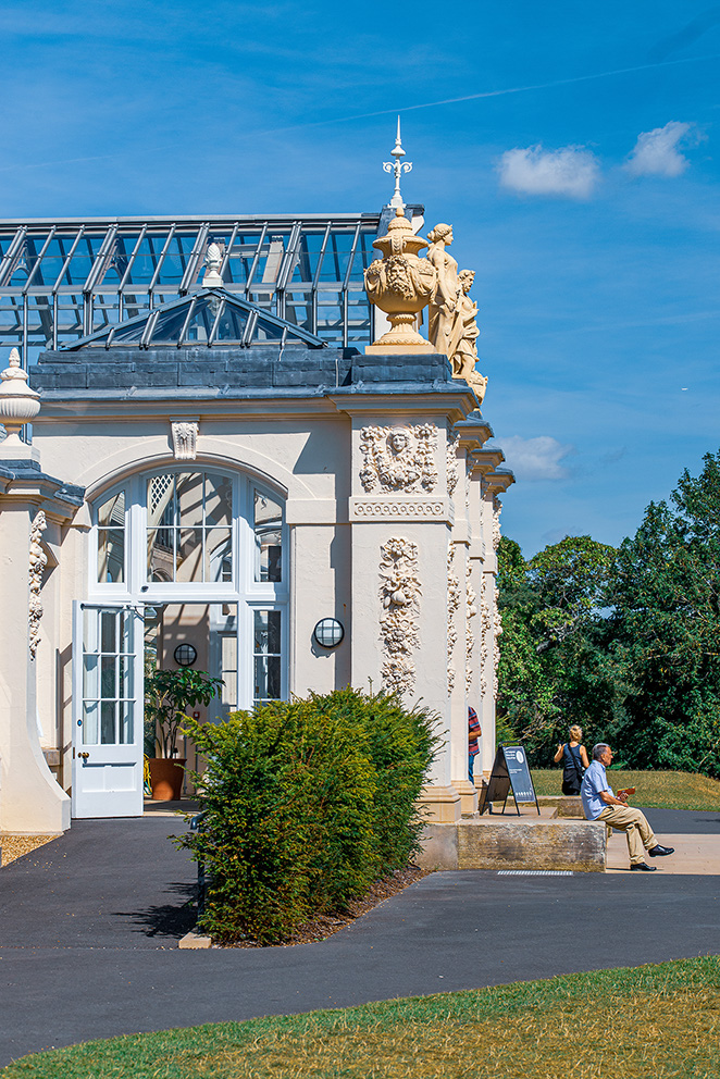 The image depicts a historic building with ornate architectural details, including statues and decorative elements. The structure features large glass windows and a glass roof, suggesting it might be a conservatory or greenhouse. Outside, there is a well-maintained garden with a paved walkway. Two people are visible in the scene: one sitting on a bench and another standing near a sign. The sky is clear with a few clouds, indicating a bright and sunny day.