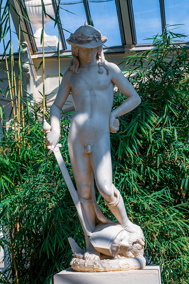 The image depicts a classical statue of a young man standing on a shell, often identified as a representation of Neptune or a sea deity. The statue is made of white marble and features intricate details, including the figure's pose and the shell base. The figure is adorned with a wreath on his head and holds a trident in his right hand. The statue is set outdoors, surrounded by greenery, with a modern glass structure in the background.