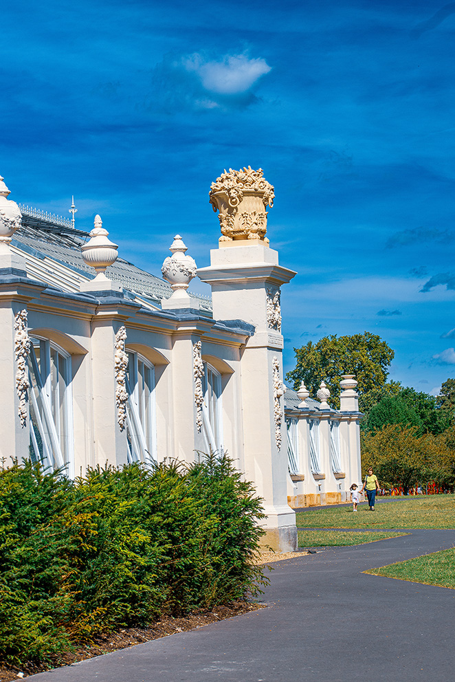 The image depicts a section of an ornate, white building with classical architectural elements, including columns and decorative urns. The structure is set against a bright blue sky with a few clouds. There is a paved walkway in the foreground, flanked by greenery and trees, with a person and a child walking along it.