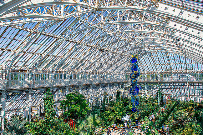 The image depicts the interior of a large greenhouse or conservatory with a high, arched, glass-and-metal ceiling. The structure allows ample natural light to illuminate the lush, diverse plant life below. A prominent, colorful sculpture of a double helix DNA strand hangs from the ceiling, adding an artistic element to the natural surroundings. People can be seen walking and observing the plants, indicating that this is a public space, likely a botanical garden or similar attraction.